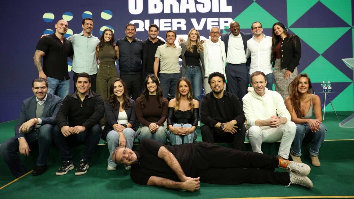 Fotografia em plano aberto de um grande grupo de profissionais de comunicação e esportes reunidos em um palco com piso verde e fundo azul, posando juntos em duas fileiras principais com um homem deitado no chão à frente