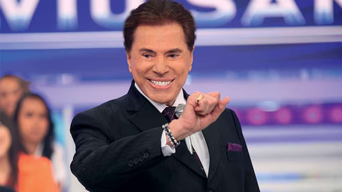 Fotografia em plano médio do apresentador Silvio Santos sorrindo de terno escuro e gesticulando com a mão esquerda à frente em seu programa de televisão