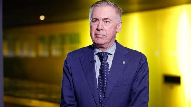 Fotografia em plano médio do técnico Carlo Ancelotti vestindo terno azul e gravata com uma expressão focada diante de um fundo iluminado