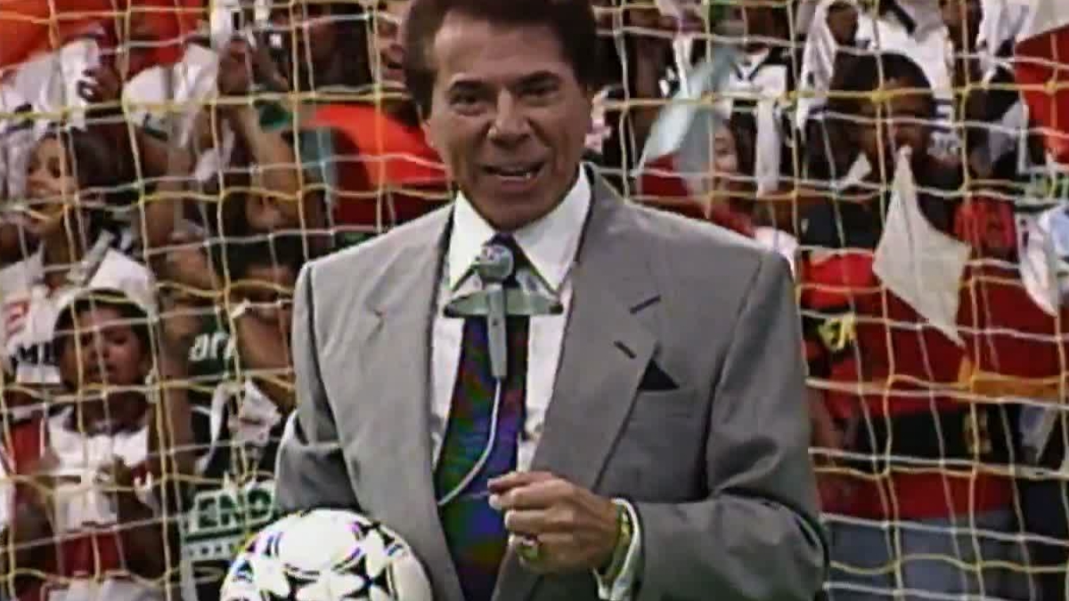 Fotografia de arquivo do apresentador Silvio Santos sorrindo de terno cinza e gravata escura enquanto segura uma bola de futebol à frente de uma rede de gol com pessoas ao fundo