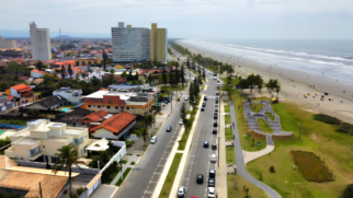Estrutura Urbana e Construções Do lado esquerdo, ergue-se o tecido urbano composto por casas residenciais de telhados cerâmicos em tons de laranja e marrom, misturadas a edifícios comerciais de médio porte. Mais ao fundo, destacam-se alguns prédios residenciais altos e de arquitetura retangular branca e amarela. Uma avenida de pista dupla, dividida por um canteiro central gramado com coqueiros e postes de iluminação, corta verticalmente o centro da cena. Carros de cores variadas transitam ou estão estacionados ao longo das vias. Área de Lazer e Orla À direita da avenida, estende-se uma ampla faixa de vegetação rústica e gramados bem cuidados. Nessa área de lazer, destaca-se uma pista de pump track (circuito sinuoso e asfaltado para bicicletas, skates ou patins) circundada por uma calçada cinza para pedestres. Pequenas figuras humanas caminham pelas passarelas e pela ciclovia adjacente. Praia e Mar Na extremidade direita, uma extensa e larga faixa de areia clara e firme faz a transição para o oceano. O mar apresenta águas de tom esverdeado com linhas consecutivas de ondas e espuma branca que quebram suavemente na areia. Pequenos pontos coloridos espalhados pela praia indicam a presença de banhistas, guarda-sóis e quiosques sob o céu claro e parcialmente nublado.
