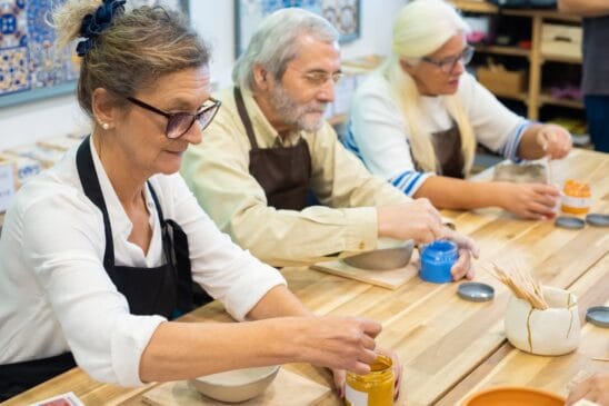 Três idosos participam de uma oficina de cerâmica enquanto pintam peças artesanais em uma mesa de madeira.