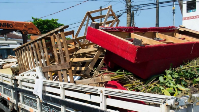 Esta imagem registra a carroceria de um caminhão de zeladoria urbana carregada com diversos materiais volumosos e resíduos descartados. No topo da carga, sobressai a estrutura de madeira de um sofá com estofamento vermelho vibrante, acompanhada por estrados de cama e restos de móveis desmontados. Sob essas estruturas, nota-se uma quantidade significativa de resíduos orgânicos, como galhos, folhas e restos de podas. O caminhão é branco e conta com um braço mecânico laranja na lateral, utilizado para o içamento de objetos pesados. A cena ocorre em uma via pública durante o dia, com fiações elétricas e construções simples ao fundo, reforçando o contexto de uma operação de limpeza e manutenção da infraestrutura da cidade.