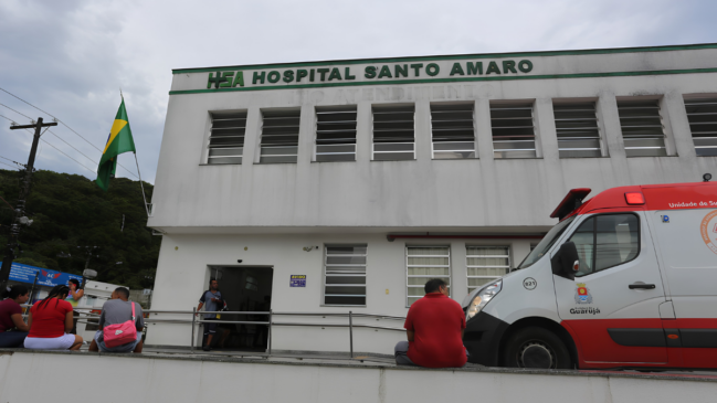 A fotografia mostra a fachada do Hospital Santo Amaro, em Guarujá, sob um céu nublado. O prédio é uma construção de dois andares com paredes brancas. No topo, há uma faixa horizontal verde com o logotipo "HSA" e o nome "HOSPITAL SANTO AMARO" em letras garrafais verdes. Além disso, do lado esquerdo da fachada, uma bandeira do Brasil está hasteada em um mastro preso à parede. Em primeiro plano, à direita, aparece parte de uma ambulância branca com detalhes em vermelho, onde se lê "Prefeitura de Guarujá" e o brasão do município na porta lateral. Nesse sentido, o veículo reforça a presença do serviço público na unidade. No centro e à esquerda da imagem, algumas pessoas estão sentadas em uma mureta de cimento em frente à entrada do hospital, sugerindo espera por atendimento. Dessa forma, a cena captura o cotidiano da instituição de saúde. Portanto, a imagem contextualiza o local que recebeu a notificação oficial da administração municipal. Desta maneira, a composição fotográfica evidencia tanto a infraestrutura hospitalar quanto a integração com os serviços de emergência da cidade de Guarujá.