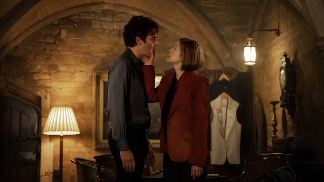 Uma fotografia de plano médio e horizontal captura uma interação tensa e íntima entre um casal, no interior de uma sala de estar com decoração rústica e sofisticada, sob iluminação âmbar suave. Elementos em Destaque A Mulher (Lado Direito): Uma mulher de cabelos loiros curtos (corte bob), na altura do queixo, está de pé e ligeiramente voltada para a esquerda. Ela veste um blazer de linho vermelho-terracota estruturado sobre uma blusa escura e calça preta. Seu braço direito está levantado e estendido, com a mão direita tocando delicadamente o queixo e a face inferior do homem, mantendo um olhar fixo e intenso nos olhos dele. Ela usa um anel dourado simples na mão esquerda. O Homem (Lado Esquerdo): Um homem de cabelos pretos e curtos está de pé e de frente para a mulher, aceitando o toque dela. Ele veste uma camisa de manga longa de cetim azul-petróleo (cinza-azulado escuro) e calça preta. Ele está com a cabeça ligeiramente inclinada para a direita, correspondendo ao contato visual da mulher. O Cenário (Ao Fundo): O ambiente tem paredes de pedra rústica e um teto arqueado de tijolos expostos, sugerindo uma adega ou construção antiga. No fundo, entre o casal, há um biombo de madeira escura dobrado e, pendurados nele, alguns cabides com roupas (uma jaqueta preta e um colete branco). À esquerda, vê-se um abajur de mesa com cúpula plissada bege aceso, sobre um móvel de madeira. À direita, um candeeiro de parede de ferro forjado no estilo lanterna está aceso, ao lado de um relógio de parede antigo de pêndulo. Iluminação, Cores e Clima Efeitos e Luz: A luz é quente e difusa, vinda principalmente do abajur e do candeeiro, projetando sombras suaves e destacando as texturas da pedra e do tecido do blazer. Cores: A paleta é dominada por tons terrosos e quentes (vermelho-terracota, âmbar, marrom, dourado), contrastando com o azul-escuro e preto das vestimentas do homem e a camisa da mulher. Ambiente: A atmosfera é envolvente, dramática e carrega uma tensão emocional palpável entre os dois personagens.