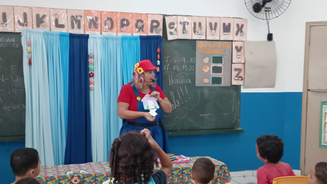 A imagem registra uma cena em plano médio capturada dentro de uma sala de aula de educação infantil. Uma educadora fantasiada realiza uma atividade lúdica de contação de histórias para um grupo de crianças pequenas. Elementos Principais (Primeiro Plano e Centro) A Educadora: Centralizada na imagem, uma mulher de pele clara e cabelos loiros com mechas coloridas aparece de pé, voltada levemente para a sua direita. Ela usa um figurino alegre composto por uma camiseta vermelha, um macacão jeans de saia rodada azul-escura e um quepe vermelho adornado com girassóis artificiais amarelos na lateral. Ela utiliza um microfone auricular (headset) preto e segura com as duas mãos um pequeno cenário ou fantoche de papel em formato de casa com uma figura branca na frente. As Crianças: Sentadas de costas ou de perfil para a câmera, na metade inferior da imagem, as crianças observam atentamente a apresentação. Em destaque no centro, está uma menina com cabelos cacheados escuros presos em duas marias-chiquinhas, que levanta a mão direita. À esquerda, um menino de cabelos raspados acompanha a atividade, e à direita, outro menino de camiseta vermelha observa a professora. Uma mesa baixa coberta com uma toalha estampada e colorida está posicionada entre as crianças e a contadora. Elementos Secundários (Plano de Fundo) A Lousa e os Cartazes: Atrás da professora, estende-se uma grande lousa verde escolar fixa na parede, que exibe anotações em giz branco com números de 7 a 20 e a palavra "HISTÓRIA" escrita no centro. Acima e ao lado da lousa, há uma faixa com cartazes do alfabeto ilustrado, exibindo letras maiúsculas de "I" a "Z" acompanhadas de desenhos e nomes de animais ou objetos. Um cartaz colorido com o título "QUANTOS SOMOS?" está fixado na lousa. A Decoração da Sala: Do lado esquerdo da professora, cobrindo parte da lousa, há uma cortina de tecido em tiras verticais com três tons de azul (claro, médio e escuro), decorada com pequenos enfeites florais pendurados. A parede da sala possui uma divisão de cores, sendo a metade superior branca e a metade inferior pintada em azul-vivo. No canto superior direito, um ventilador de parede preto está visível próximo a uma porta de madeira clara.