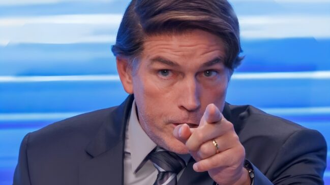 A imagem apresenta um plano fechado do ator Jim Caviezel, centralizado, em uma postura que transmite autoridade e confronto. Ele é um homem de pele clara, com cabelos castanhos curtos, penteados para o lado e levemente grisalhos nas têmporas. Sua expressão facial é severa e focada, com sobrancelhas levemente franzidas e olhos fixos diretamente para a frente. O ator veste um traje formal composto por um paletó escuro, camisa social clara e uma gravata com listras diagonais em tons de cinza e preto. O elemento de maior destaque na composição é sua mão direita, que está estendida em direção ao espectador com o dedo indicador apontado diretamente para a câmera, criando um efeito de perspectiva. Na mão que aponta, é possível visualizar uma aliança dourada no dedo anelar. O fundo da imagem é composto por linhas horizontais em tons de azul vibrante e branco, que estão desfocadas, sugerindo um ambiente de estúdio de televisão ou cenário de entrevista. A iluminação é frontal e nítida, destacando os detalhes do rosto e as texturas das roupas, enquanto a profundidade de campo reduzida mantém o foco estritamente no gesto e na expressão do ator.