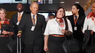 A imagem colorida mostra um grupo de seis profissionais da aviação civil, provavelmente tripulantes de cabine e pilotos da companhia aérea GOL, caminhando juntos em um ambiente aeroportuário. Eles estão enfileirados lado a lado, em movimento em direção à câmera, e exibem expressões sorridentes e profissionais. Detalhes das Pessoas (da esquerda para a direita): Primeira mulher: Tem cabelos loiros presos, sorri e veste um uniforme preto com um lenço avermelhado amarrado no pescoço. Ela carrega uma bolsa preta de mão. Primeiro homem (ao fundo): É um homem negro de barba, sorrindo amplamente com os dentes à mostra. Ele usa um terno preto, camisa branca, gravata laranja e um crachá de identificação pendurado no pescoço por um cordão também laranja. Segundo homem (em destaque): É um homem de pele clara e cabelos grisalhos, com aparência mais madura, representando a figura de um comandante/piloto. Ele veste terno preto, camisa branca, gravata cinza e usa o cordão laranja com crachá. Com a mão direita, ele puxa uma mala de rodinhas preta. Segunda mulher (no centro): Tem cabelos longos e castanhos, usa óculos de grau com armação fina e sorri. Ela veste uma camisa social branca de mangas curtas, saia preta com cinto laranja e o lenço avermelhado no pescoço. Ela tem tatuagens visíveis no braço direito e carrega uma bolsa preta no braço esquerdo. Terceira mulher (ao fundo): Tem cabelos pretos presos em um coque, pele clara e olha sorrindo para a direita. Veste um terno preto com camisa branca, gravata escura e o cordão laranja com crachá. Quarta mulher (extrema direita): É uma mulher negra de cabelos curtíssimos ou raspados, sorrindo e usando batom vermelho. Ela veste camisa branca, o lenço avermelhado característico no pescoço e segura um paletó preto dobrado no braço. Cenário e Elementos de Fundo: O grupo caminha de forma alinhada e confiante. Todos os integrantes da equipe utilizam crachás com foto posicionados na altura do peito ou da cintura. Ao fundo e acima deles, é possível ver de forma desfocada um painel eletrônico de voos preto, exibindo informações laranjas de horários e status de partidas e chegadas ("Previsto"). A iluminação do local é típica de um saguão ou área de embarque de aeroporto moderno.