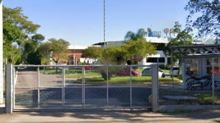 A imagem registra uma fotografia externa em plano médio de uma das sedes da empresa Dell Technologies no Brasil, capturada a partir da área externa da propriedade em um dia ensolarado. Elementos Principais (Primeiro Plano) O Portão e a Entrada: O primeiro plano é dominado por um grande portão de correr metálico na cor cinza-claro, composto por grades quadriculadas finas. Ele está fechado e é sustentado por pilares de concreto cinza nas extremidades. A Guarita: À direita do portão, há uma guarita de segurança cinza com janelas de vidro. Logo em frente à guarita, do lado de dentro do cercado de proteção, encontra-se estacionada uma motocicleta de cor escura posicionada de perfil. Elementos Secundários (Plano de Fundo) O Complexo e o Letreiro: Atrás do portão, ergue-se o complexo empresarial de estrutura horizontal e paredes brancas. No topo do edifício principal, destaca-se um letreiro azul com o logotipo da marca "DELL". Próximo ao letreiro, há antenas de comunicação instaladas no teto. O Pátio Interno e a Vegetação: Uma via pavimentada interna estende-se do lado esquerdo em direção ao fundo da propriedade. O pátio conta com extensas áreas de gramado verde bem cuidado, canteiros redondos e diversas árvores de médio porte com copas folheadas que sombreiam parcialmente o espaço. Alguns veículos de passeio, incluindo um carro vermelho e outro prata, aparecem estacionados entre os jardins ao fundo. O Céu: A parte superior da composição mostra um céu azul limpo, sem nuvens, indicando iluminação natural direta e intensa sobre todo o complexo.