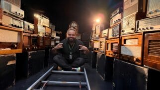 A imagem mostra um homem sorridente sentado de pernas cruzadas no centro de um cenário cinematográfico, posicionado sobre trilhos de câmera (travelling). Ele veste uma camiseta preta, usa um crachá no pescoço e faz gestos com as mãos enquanto está ladeado por grandes pilhas de rádios antigos de diversos modelos e épocas, muitos com acabamento em madeira e mostradores analógicos. O ambiente possui uma iluminação dramática de estúdio, com um foco de luz quente ao fundo que cria um efeito de brilho (lens flare) e destaca as texturas dos aparelhos vintage empilhados sobre caixas pretas, sugerindo os bastidores de uma produção audiovisual relacionada à história do rádio.
