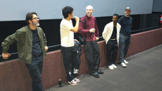 Esta fotografia colorida registra, em plano médio e ângulo ligeiramente elevado, cinco homens alinhados em frente à tela de uma sala de cinema. O grupo participa de um debate ou apresentação, possivelmente no Cine Roxy. Da esquerda para a direita: Um homem de óculos e barba grisalha veste uma jaqueta bomber verde-oliva sobre camiseta preta, mantendo uma postura de observação. Um jovem de camiseta branca segura um microfone e fala em direção aos outros integrantes do grupo. No centro, um jovem de pele clara e cabeça raspada veste um blusão bordô e calças largas escuras, ouvindo atentamente. Ao lado dele, um jovem negro usa uma jaqueta branca aberta sobre uma camiseta preta e calças escuras. Na extremidade direita, um homem de camiseta preta, boné e óculos observa a interação de braços cruzados. Abaixo da tela de cinema, uma mureta revestida em carpete marrom serve de apoio para alguns dos presentes. O ambiente possui iluminação artificial direta, típica de salas de exibição, e o chão é escuro. Uma lata de bebida aparece no rodapé da imagem, próximo ao homem de camiseta branca.