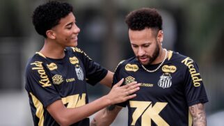 Esta imagem captura um momento de interação entre dois jogadores de futebol durante um treinamento no CT Rei Pelé. À esquerda, o jovem Robinho Jr. aparece sorrindo e olhando para o companheiro, enquanto coloca a mão direita de forma amigável no peito de Neymar. À direita, o camisa 10 do Santos está com o olhar baixo, demonstrando uma expressão mais séria ou reflexiva. Ambos vestem o uniforme de treino oficial do Santos FC, composto por uma camisa preta com detalhes em dourado e os logotipos dos patrocinadores (como Kicaldo e Canção) em evidência. Neymar utiliza acessórios marcantes, como um colar de pérolas, brincos e um anel na mão esquerda. A fotografia possui um fundo desfocado, destacando os atletas em primeiro plano, e ilustra visualmente a relação de "padrinho e afilhado" profissional que o seu texto menciona, capturando a reconciliação após o desentendimento ocorrido.