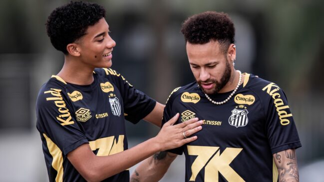 Esta imagem captura um momento de interação entre dois jogadores de futebol durante um treinamento no CT Rei Pelé. À esquerda, o jovem Robinho Jr. aparece sorrindo e olhando para o companheiro, enquanto coloca a mão direita de forma amigável no peito de Neymar. À direita, o camisa 10 do Santos está com o olhar baixo, demonstrando uma expressão mais séria ou reflexiva. Ambos vestem o uniforme de treino oficial do Santos FC, composto por uma camisa preta com detalhes em dourado e os logotipos dos patrocinadores (como Kicaldo e Canção) em evidência. Neymar utiliza acessórios marcantes, como um colar de pérolas, brincos e um anel na mão esquerda. A fotografia possui um fundo desfocado, destacando os atletas em primeiro plano, e ilustra visualmente a relação de "padrinho e afilhado" profissional que o seu texto menciona, capturando a reconciliação após o desentendimento ocorrido.