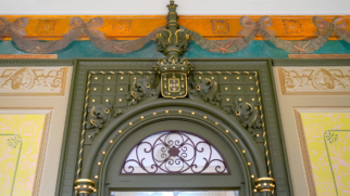 A imagem mostra um detalhe arquitetônico em close-up da parte superior de um portal antigo e ricamente decorado, evidenciando elementos clássicos e restauração artística. O Portal Central No centro da imagem, destaca-se a estrutura em arco de uma porta, pintada em tom verde-oliva escuro. O arco é adornado com pequenas esferas douradas em relevo ao longo de sua curvatura. Acima do arco, há um painel retangular quadriculado, também em verde, decorado com pontos dourados em cada quadrado e esculturas ornamentais de folhagens nas laterais. No topo central desse conjunto, repousa um brasão de armas clássico (semelhante ao escudo de Portugal), contornado em dourado e encimado por uma coroa estilizada da mesma cor. Acima do brasão, uma agulha ou pináculo vertical verde e dourado estende-se até o teto. Abaixo do arco principal, há uma bandeira de vidro com uma grade de ferro batido em arabescos pretos. Detalhes das Paredes Laterais Nas laterais esquerda e direita do portal verde, as paredes exibem painéis verticais com molduras finas. O interior dessas molduras apresenta uma pintura ornamental com padronagem floral em tons vibrantes de amarelo-claro e detalhes sutis em verde e rosa. Acima desses painéis amarelos, há uma faixa horizontal em tom bege texturizado com frisos decorativos em relevo, que simulam ramos e folhas esculpidas. O Forro Superior Na parte mais alta da imagem, rente ao teto branco, estende-se um amplo barrado pictórico horizontal. Esse barrado combina uma base azul-turquesa na parte inferior com uma faixa amarela-ouro ou dourada na parte superior. Sobre essa faixa dourada, há uma pintura detalhada que simula um tecido em formato de guirlanda ou laço contínuo, entrelaçado com cordões e pingentes em tons de cinza e marrom, conferindo profundidade tridimensional à obra de arte.