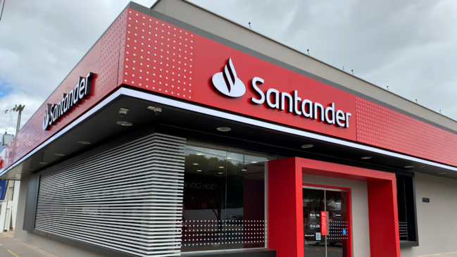 A imagem mostra uma fotografia em plano médio e ângulo diagonal da fachada externa de uma agência bancária do Santander, registrada sob um céu nublado e acinzentado. Estrutura da Fachada A Platibanda Superior (Cobertura): O topo do edifício apresenta uma estrutura arquitetônica moderna em formato de "L", que contorna a esquina da agência. Essa platibanda é revestida por painéis na cor vermelha viva, decorados com um padrão de pequenos pontos brancos dispostos em linhas verticais nas extremidades. O Logotipo da Marca: Fixado em destaque na parte central e lateral esquerda do painel vermelho, encontra-se o logotipo oficial do banco em relevo tridimensional. À esquerda, há o símbolo da chama estilizada na cor branca com contornos escuros. À direita do símbolo, está escrita a palavra "Santander" em letras brancas com tipografia arredondada característica. A Base e o Teto Técnico: Abaixo do painel vermelho, há uma moldura horizontal preta que abriga lâmpadas embutidas e câmeras de segurança esféricas (domes) voltadas para a calçada. Área Térrea e Entrada As Vidraças e Brises: A fachada frontal é composta por grandes painéis de vidro transparente que revelam o interior iluminado da agência. Na lateral esquerda, protegendo parte da vidraça, há uma estrutura de brises horizontais de alumínio branco (grades de sombreamento). No vidro, há uma inscrição interna parcial onde se lê: "POR VOCÊ HOJE?". A Entrada Principal: À direita, o portal de acesso ao banco é emoldurado por uma grande estrutura geométrica e saltada também na cor vermelha. As portas de vidro de correr contam com adesivos de sinalização, incluindo uma placa vermelha com informações de horários de atendimento e um símbolo azul de acessibilidade para cadeirantes. O Entorno: As paredes das extremidades do prédio possuem pintura texturizada em tom cinza-escuro. No canto esquerdo, nota-se uma calçada estreita e um poste de energia elétrica ao fundo, sob o tempo encoberto.