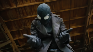 Uma imagem colorida na horizontal e em plano médio, capturada de um ângulo levemente contra-plongée (de baixo para cima), foca no herói Spider-Noir. Na imagem, o personagem aparece centralizado, vestido com seu traje característico inspirado no gênero noir dos anos 1930. O herói usa uma máscara preta de tecido com textura sutil e costuras visíveis na parte superior, equipada com grandes lentes oculares brancas, ovais e saltadas, que possuem armações pretas grossas. Ele veste um sobretudo de gola larga em tom cinza-escuro ou marrom-envelhecido sobre uma roupa preta justa, complementado por luvas escuras de couro em ambas as mãos. Sua postura é dinâmica: o torso está levemente inclinado para a frente enquanto ele estende os braços em direção à câmera. Suas mãos estão posicionadas no clássico gesto de disparo de teias do Homem-Aranha, com os dedos médios e anelares recolhidos contra a palma e os dedos polegar, indicador e mínimo estendidos. O fundo da cena é composto por vigas e ripas de madeira rústica em tons de marrom, sugerindo o teto ou o interior de um sótão ou galpão antigo, com iluminação suave focada no personagem.