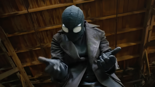 Uma imagem colorida na horizontal e em plano médio, capturada de um ângulo levemente contra-plongée (de baixo para cima), foca no herói Spider-Noir. Na imagem, o personagem aparece centralizado, vestido com seu traje característico inspirado no gênero noir dos anos 1930. O herói usa uma máscara preta de tecido com textura sutil e costuras visíveis na parte superior, equipada com grandes lentes oculares brancas, ovais e saltadas, que possuem armações pretas grossas. Ele veste um sobretudo de gola larga em tom cinza-escuro ou marrom-envelhecido sobre uma roupa preta justa, complementado por luvas escuras de couro em ambas as mãos. Sua postura é dinâmica: o torso está levemente inclinado para a frente enquanto ele estende os braços em direção à câmera. Suas mãos estão posicionadas no clássico gesto de disparo de teias do Homem-Aranha, com os dedos médios e anelares recolhidos contra a palma e os dedos polegar, indicador e mínimo estendidos. O fundo da cena é composto por vigas e ripas de madeira rústica em tons de marrom, sugerindo o teto ou o interior de um sótão ou galpão antigo, com iluminação suave focada no personagem.