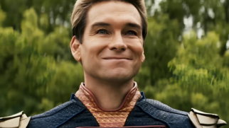 A imagem apresenta um plano médio, do peito para cima, do personagem Capitão Pátria (interpretado pelo ator Antony Starr), da série The Boys. O personagem é um homem branco de meia-idade, com cabelos loiros curtos, penteados para trás e impecavelmente alinhados. Ele exibe um sorriso fechado e contido, que pressiona os lábios e cria rugas de expressão ao redor dos olhos azuis, transmitindo uma aparência de satisfação irônica ou condescendente. Sua pele é clara e apresenta marcas naturais de expressão na testa e nas bochechas. Ele veste seu traje de super-herói azul-marinho com textura de escamas. A gola é alta, em um tom dourado envelhecido com padrões geométricos, e possui um acabamento bordô. Nos ombros, destacam-se as ombreiras metálicas douradas que fazem parte da composição clássica de seu uniforme. O fundo da imagem está desfocado, revelando uma densa folhagem verde de árvores sob uma iluminação natural clara, o que sugere um ambiente externo durante o dia. O foco permanece inteiramente na expressão facial do personagem.