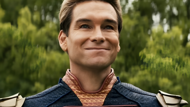 A imagem apresenta um plano médio, do peito para cima, do personagem Capitão Pátria (interpretado pelo ator Antony Starr), da série The Boys. O personagem é um homem branco de meia-idade, com cabelos loiros curtos, penteados para trás e impecavelmente alinhados. Ele exibe um sorriso fechado e contido, que pressiona os lábios e cria rugas de expressão ao redor dos olhos azuis, transmitindo uma aparência de satisfação irônica ou condescendente. Sua pele é clara e apresenta marcas naturais de expressão na testa e nas bochechas. Ele veste seu traje de super-herói azul-marinho com textura de escamas. A gola é alta, em um tom dourado envelhecido com padrões geométricos, e possui um acabamento bordô. Nos ombros, destacam-se as ombreiras metálicas douradas que fazem parte da composição clássica de seu uniforme. O fundo da imagem está desfocado, revelando uma densa folhagem verde de árvores sob uma iluminação natural clara, o que sugere um ambiente externo durante o dia. O foco permanece inteiramente na expressão facial do personagem.