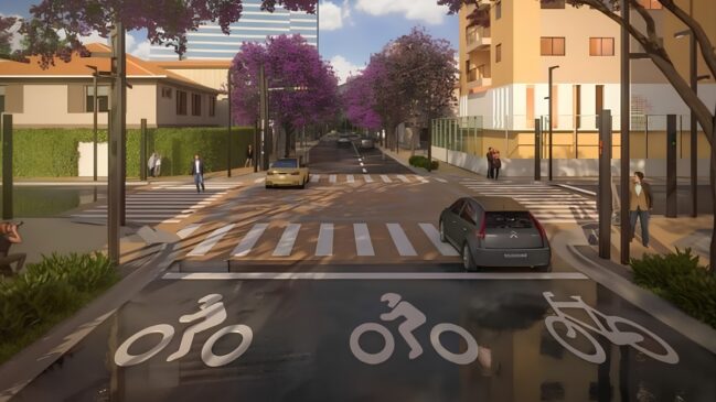 A imagem apresenta uma projeção arquitetônica tridimensional (renderização) que ilustra o projeto de reurbanização de uma via urbana, com foco em acessibilidade, paisagismo moderno e organização do trânsito. A cena é retratada a partir de uma perspectiva central, ao nível da rua. Primeiro Plano e Ciclovia Na parte inferior da imagem, em primeiro plano, destaca-se um trecho de ciclovia com pavimento asfáltico escuro e liso. Três grandes pictogramas brancos pintados no chão indicam a exclusividade para ciclistas: os dois primeiros mostram silhuetas estilizadas de pessoas pedalando e o terceiro exibe apenas o desenho de uma bicicleta. Uma linha branca contínua delimita o fim dessa faixa. O Cruzamento e os Veículos Logo após a ciclovia, há um cruzamento viário estruturado com faixas de pedestres sinalizadas em branco. O piso do cruzamento apresenta uma tonalidade marrom-clara, sugerindo o uso de blocos intertravados de concreto (pavers). Dois carros estão inseridos na via: À direita: Um veículo moderno do modelo hatchback, na cor cinza-escura, está posicionado de costas, avançando em direção ao cruzamento. Ao centro: Um carro de passeio na cor amarela ou dourada trafega no sentido oposto, mais ao fundo da via principal. Pedestres e Infraestrutura Urbana Diversas figuras humanas estilizadas estão distribuídas pelas calçadas para simular o uso do espaço público: No canto inferior esquerdo, um homem agachado parece registrar a cena. Próximo à faixa de pedestres da esquerda, um homem de terno caminha em direção à via, enquanto um casal conversa mais ao fundo, perto de uma cerca viva alta e verde. Na calçada da direita, um pedestre caminha em primeiro plano e, mais atrás, um casal anda abraçado perto de um edifício residencial de cor amarela-clara com varandas. Postes de iluminação pública modernos, esguios e na cor preta estão posicionados estrategicamente ao longo das calçadas em ambos os lados da rua. Nota-se a total ausência de fiação aérea. Paisagismo e Elementos ao Fundo O projeto paisagístico é um dos grandes destaques da imagem. Árvores de médio porte com copas densas e repletas de flores na cor roxa ou jambo (características de ipês) margeiam os dois lados da avenida, estendendo-se em direção ao fundo da perspectiva. Canteiros com vegetação rasteira verde decoram as esquinas em primeiro plano. Ao fundo, a rua continua em linha reta com mais veículos distantes, sob um céu azul-claro com nuvens brancas esparsas. No horizonte, ergue-se a silhueta de um edifício comercial alto e moderno com fachada espelhada em tons azulados.