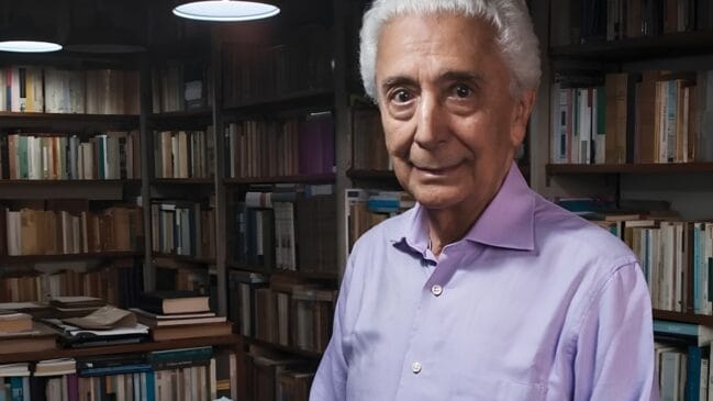 A imagem apresenta o historiador Fernando Antonio Novais em um ambiente que reforça sua trajetória intelectual, posicionado em primeiro plano e levemente à direita da composição. Ele possui cabelos brancos e curtos, veste uma camisa social de cor lilás clara e exibe uma expressão serena, com um leve sorriso e olhar direto para a câmera. Ao fundo, diversas estantes repletas de livros ocupam quase todo o cenário, criando uma profundidade de campo que destaca a figura do professor em relação à sua biblioteca pessoal ou acadêmica. A iluminação é suave e provém de luminárias no teto, que lançam focos de luz sobre as prateleiras à esquerda, enquanto o restante do ambiente mantém uma tonalidade mais sóbria e acolhedora, típica de um escritório de estudos ou arquivo histórico.