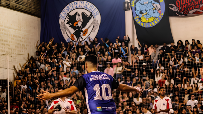 A imagem retrata a atmosfera vibrante de uma partida de handebol durante os Jogos da Unisanta, capturando um jogador da equipe de Engenharia de costas, vestindo a camisa azul de número 10, com os braços abertos em direção à torcida. Ao fundo, uma arquibancada lotada exibe o entusiasmo dos estudantes, que estão em pé e vibrando atrás de uma rede de proteção, sob grandes bandeiras que representam os cursos de Engenharia e Odontologia da instituição. A cena foca na interação entre o atleta e o público, evidenciando a paixão e a tradição esportiva que envolvem essa competição universitária na Baixada Santista.