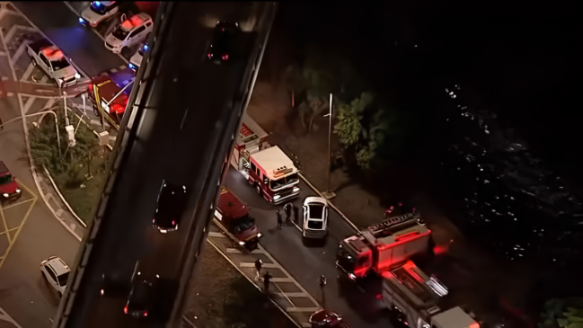 Uma vista aérea noturna captura a movimentação intensa de viaturas de emergência em uma via expressa urbana de São Paulo, logo após um grave acidente automobilístico. A imagem é registrada de um ângulo diagonal superior, oferecendo uma perspectiva ampla do local. Elementos em Destaque O Viaduto Elevado (Eixo Central-Esquerdo): Uma estrutura de viaduto corta a imagem verticalmente, ligeiramente inclinada para a esquerda. Sobre ele, poucos veículos de passeio escuros trafegam em velocidade reduzida. A pista do viaduto está parcialmente iluminada pelos postes públicos e pelos faróis dos próprios carros. A Área do Acidente e Resgate (Eixo Central e Inferior): Abaixo do viaduto, na pista local, concentra-se a força-tarefa de salvamento. Há pelo menos dois grandes caminhões do Corpo de Bombeiros com luzes de sinalização ativas, além de ambulâncias e viaturas policiais posicionadas estrategicamente para isolar a área. Um carro de passeio branco está estacionado de forma transversal no centro da pista, cercado por socorristas. O Rio e as Margens (Lado Direito): À direita da pista de resgate, nota-se uma área de vegetação escura (árvores e arbustos) que margeia o leito do Rio Tamanduateí. A superfície da água aparece escura e com reflexos sutis das luzes das viaturas, indicando a proximidade imediata do ponto onde o veículo despencou. O Acesso Viário (Canto Superior Esquerdo): No topo esquerdo, vê-se a alça de acesso e o cruzamento de pistas sinalizadas com faixas pintadas no asfalto, onde outras viaturas com girofretos ligados dão suporte ao bloqueio do tráfego. Iluminação e Clima Visual Efeitos de Luz: A cena é predominantemente escura devido ao horário noturno (madrugada), mas recebe forte iluminação artificial nas cores vermelha, azul e branca, proveniente dos girofleds e das luzes de emergência dos veículos de resgate. Essas luzes criam reflexos intensos no asfalto úmido e nas superfícies dos automóveis. Atmosfera: A composição transmite uma sensação de urgência, gravidade e organização técnica das equipes de socorro diante de uma ocorrência complexa de trânsito.