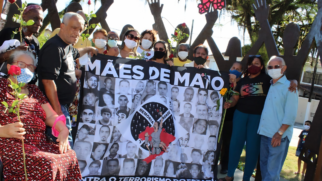 Esta imagem mostra um grupo de pessoas reunidas em um espaço público, segurando uma grande faixa do movimento Mães de Maio. O clima é de solenidade e memória.