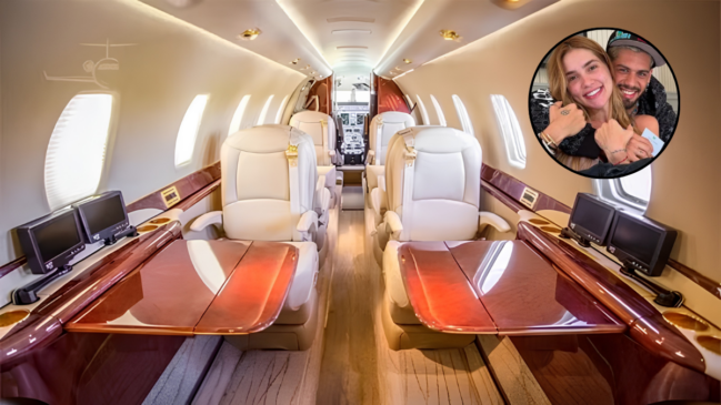 A imagem captura o interior luxuoso do jatinho particular da influenciadora Virginia Fonseca, alinhado às especificações do modelo Cessna Citation Sovereign 680 descrito no texto. A fotografia, em ângulo central de perspectiva, revela uma cabine sofisticada e extremamente confortável. Além disso, o espaço apresenta quatro poltronas executivas de couro em tom creme, organizadas em duas fileiras de frente uma para a outra, separadas por um corredor central revestido por um tapete cinza texturizado. Dessa forma, entre os assentos, mesas retráteis de madeira nobre polida estão estendidas, criando um ambiente funcional para refeições ou trabalho. A decoração utiliza amplamente acabamentos em madeira escura e brilhante nas paredes laterais, nos anteparos e nos painéis superiores, conferindo elegância ao ambiente. Nesse sentido, janelas ovais de avião ao longo da fuselagem oferecem luz natural e mostram um céu claro lá fora. O sistema de entretenimento inclui duas telas de monitor fixadas nas paredes laterais acima das mesas e outras duas telas maiores montadas nos anteparos dianteiros, próximos à porta que dá acesso à cabine de comando. Portanto, é possível visualizar o cockpit com os complexos instrumentos de voo, manetes e telas de navegação ao fundo, evidenciando a tecnologia da aeronave. Desta maneira, a iluminação interna utiliza fitas de LED no teto e luzes embutidas nos painéis laterais, criando uma atmosfera acolhedora e moderna que condiz com o padrão de vida de celebridades.