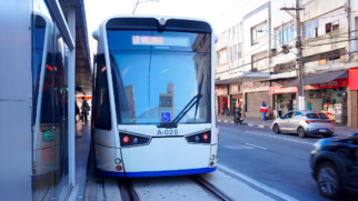 A imagem retrata o Veículo Leve Sobre Trilhos (VLT) de Santos em operação na região central da cidade. O veículo, de cor branca com detalhes em azul na base e o prefixo A-020, está parado em uma das estações da nova fase. O painel digital frontal exibe o destino "L2 VALONGO", indicando que o trem percorre a Linha 2 em direção ao Centro Histórico. Na lateral esquerda, é possível ver a estrutura da estação com portas de vidro, enquanto à direita nota-se o fluxo de veículos e o comércio local, com lojas como "Sthill Outlet" e fachadas típicas da arquitetura urbana santista. A cena ocorre durante o dia, com luz natural clara, e mostra a integração do sistema de trilhos com o trânsito de carros e pedestres na via pública.