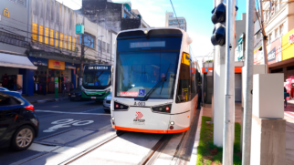 A imagem mostra o Veículo Leve Sobre Trilhos (VLT) circulando em uma via urbana de Santos durante o dia. O trem moderno possui a parte frontal predominantemente branca com detalhes em preto na área dos vidros e dos faróis, além de uma faixa laranja na base inferior, exibindo o prefixo A-003 e o logotipo da Artesp. O veículo se desloca sobre os trilhos fixados no asfalto, posicionado à direita da imagem. Do lado esquerdo, em uma faixa paralela, há um ônibus verde e branco com o letreiro indicando "Centro", além da traseira de um carro preto em movimento. O cenário ao fundo é composto pelo comércio local, calçadas, fiação aérea e postes, com um semáforo de trânsito em primeiro plano no canto direito.