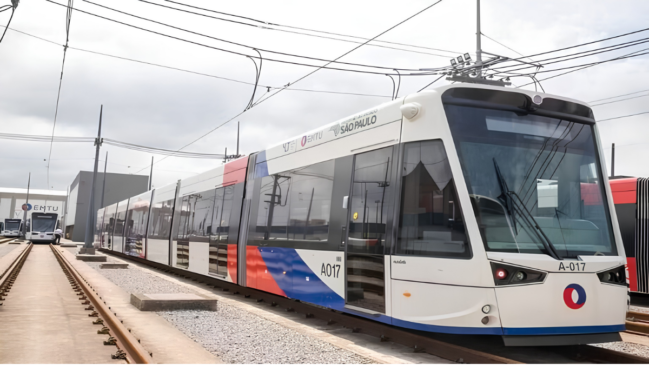 A imagem captura um moderno trem de Veículo Leve sobre Trilhos (VLT) da marca Vossloh Tramlink, estacionado em um pátio de manutenção em um dia de céu nublado, com tons predominantes de branco, azul e vermelho em sua pintura. O veículo, identificado pelo número "A017" na frente e na lateral, exibe com destaque o logotipo circular da EMTU (Empresa Metropolitana de Transportes Urbanos de São Paulo) e os textos "EMTU SÃO PAULO" e "GOVERNO DE SÃO PAULO" ao longo de sua estrutura articulada. O trem estende-se em diagonal para a esquerda, revelando múltiplos vagões e uma extensa rede de cabos de energia aérea (caténarias) suportados por postes metálicos que cortam o céu cinzento. Ele está sobre trilhos de aço apoiados em dormentes de madeira e uma camada de brita, paralelos a outras vias onde se vê, ao fundo à esquerda, outra unidade do VLT e um edifício administrativo com a identificação da EMTU, de onde uma figura solitária em uniforme de trabalho observa a cena. A composição é nítida, mostrando a infraestrutura industrial e o design funcional do transporte público metropolitano.