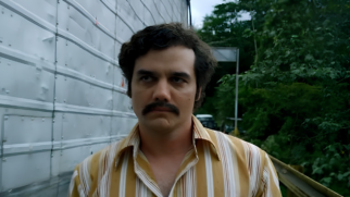 A imagem registra uma cena em plano médio do ator brasileiro Wagner Moura caracterizado como o personagem Pablo Escobar na série dramática Narcos. Elementos Principais (Primeiro Plano) O Ator: Wagner Moura aparece centralizado na imagem, enquadrado do peito para cima. Ele está com o corpo voltado para a frente, mas direciona o olhar de forma séria e compenetrada ligeiramente para o canto superior esquerdo. Características e Expressão: O ator apresenta cabelos pretos, curtos e cacheados, sobrancelhas grossas e expressivas, além de um bigode farto e escuro bem marcado acima dos lábios. Sua expressão facial é severa e pensativa, com a boca cerrada em uma linha firme. O Figurino: Ele veste uma camisa social de manga curta com colarinho aberto, caracterizada por uma estampa de listras verticais em tons de bege, ocre e branco, remetendo à estética da moda dos anos 1970 e 1980. Elementos Secundários (Plano de Fundo) As Estruturas e o Cenário: Do lado esquerdo do ator, há uma parede vertical ou barreira feita de chapas metálicas onduladas e levemente desgastadas na cor cinza-claro. A Vegetação: O lado direito do plano de fundo exibe uma área externa com vegetação densa e folhagens verdes escuras. Ao fundo, no centro da composição, é possível avistar a silhueta em desfoque de um veículo utilitário antigo estacionado em uma via de