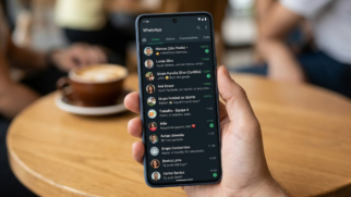 A imagem é uma fotografia em plano médio, com foco seletivo, que mostra a perspectiva de alguém segurando um smartphone em um ambiente público. Detalhes do Smartphone e da Interface No centro da imagem, uma mão masculina segura um celular com uma capa azul-escura. A tela exibe a interface do aplicativo de mensagens WhatsApp configurada em modo escuro (Dark Mode). É possível ver uma lista vertical com várias conversas e grupos, exibindo fotos de perfil circulares, nomes de contatos fictícios e trechos de mensagens em português (como "Marcos (São Paulo)", "Lucas Silva", "Grupo Família Silva (Curitiba)" e "Mãe"). Algumas conversas mostram pequenas notificações verdes com números, indicando mensagens não lidas. Elementos do Cenário O plano de fundo está fortemente desfocado (efeito bokeh), sugerindo que a cena se passa em uma cafeteria ou restaurante. À esquerda do celular, sobre uma mesa de madeira clara, nota-se uma xícara de café com leite (ou cappuccino) sobre um pires branco. Ao fundo, vultos de outras pessoas sentadas aparecem de forma sutil e sem nitidez, mantendo a atenção principal na tela do aparelho.