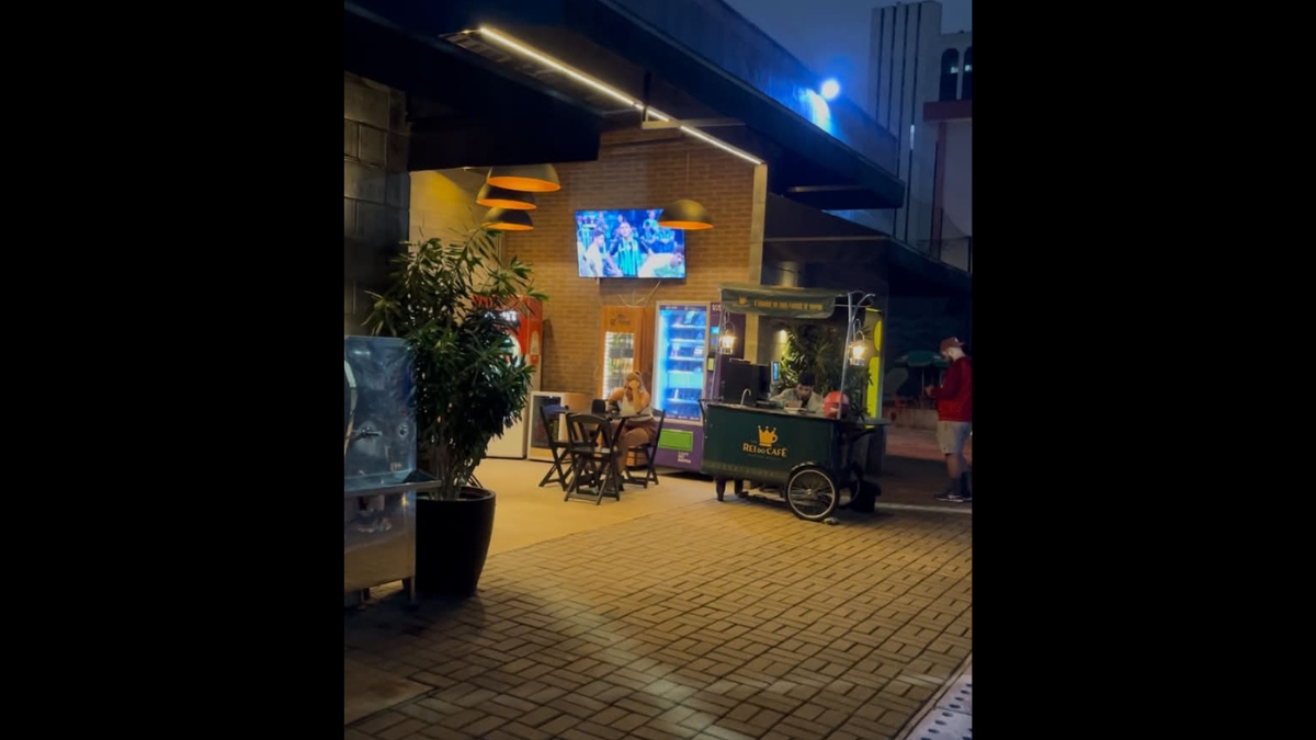 Área social noturna com mesas de madeira, um carrinho de café gourmet e uma televisão ligada em um canal de esportes.