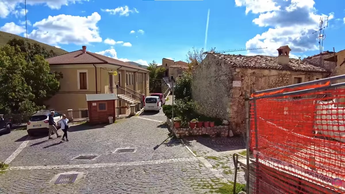 Vista de uma calçada de pedras com pedestres caminhando à esquerda e uma cerca de proteção alaranjada de obras civis à direita.