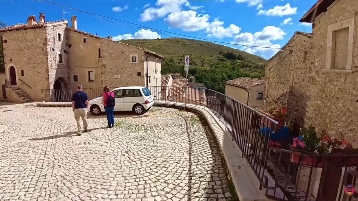 Um homem e uma mulher caminhando de costas por uma rua curva pavimentada com pedras brancas, ladeada por edifícios residenciais antigos.