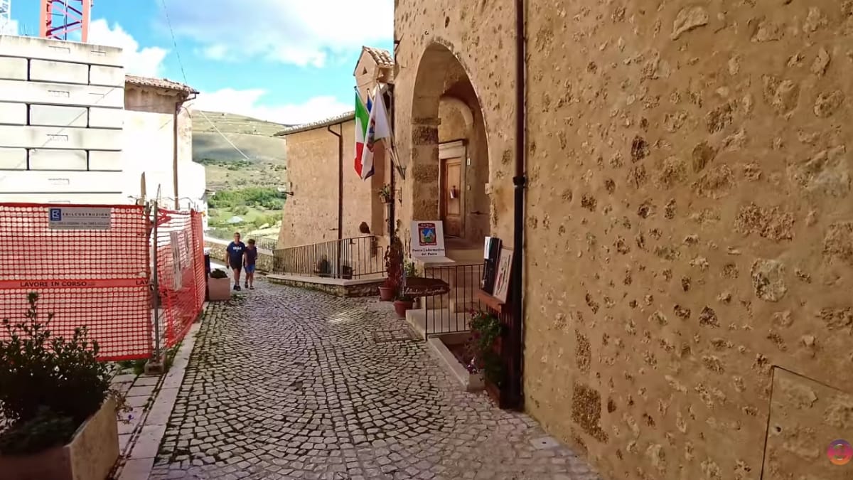Ruela de pedras que passa ao lado de um prédio de pedra com as bandeiras da Itália e da União Europeia estendidas na janela superior.