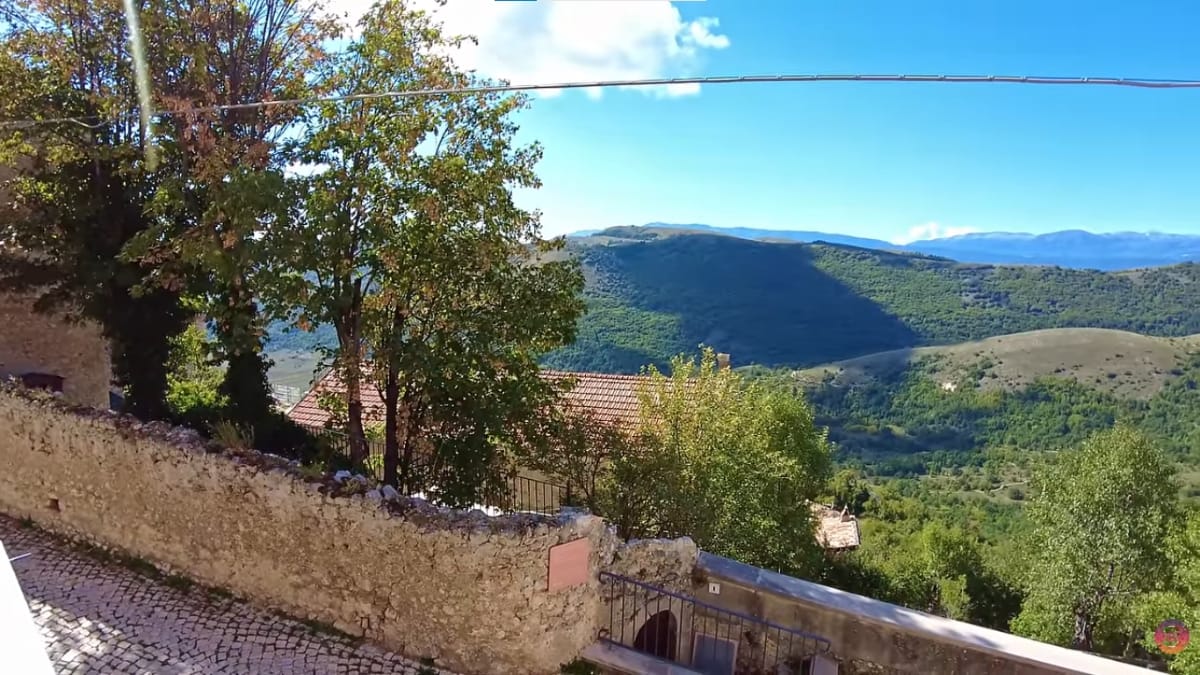 Vista a partir de uma mureta de pedra antiga mostrando uma encosta verdejante com montanhas onduladas ao fundo sob um céu azul.