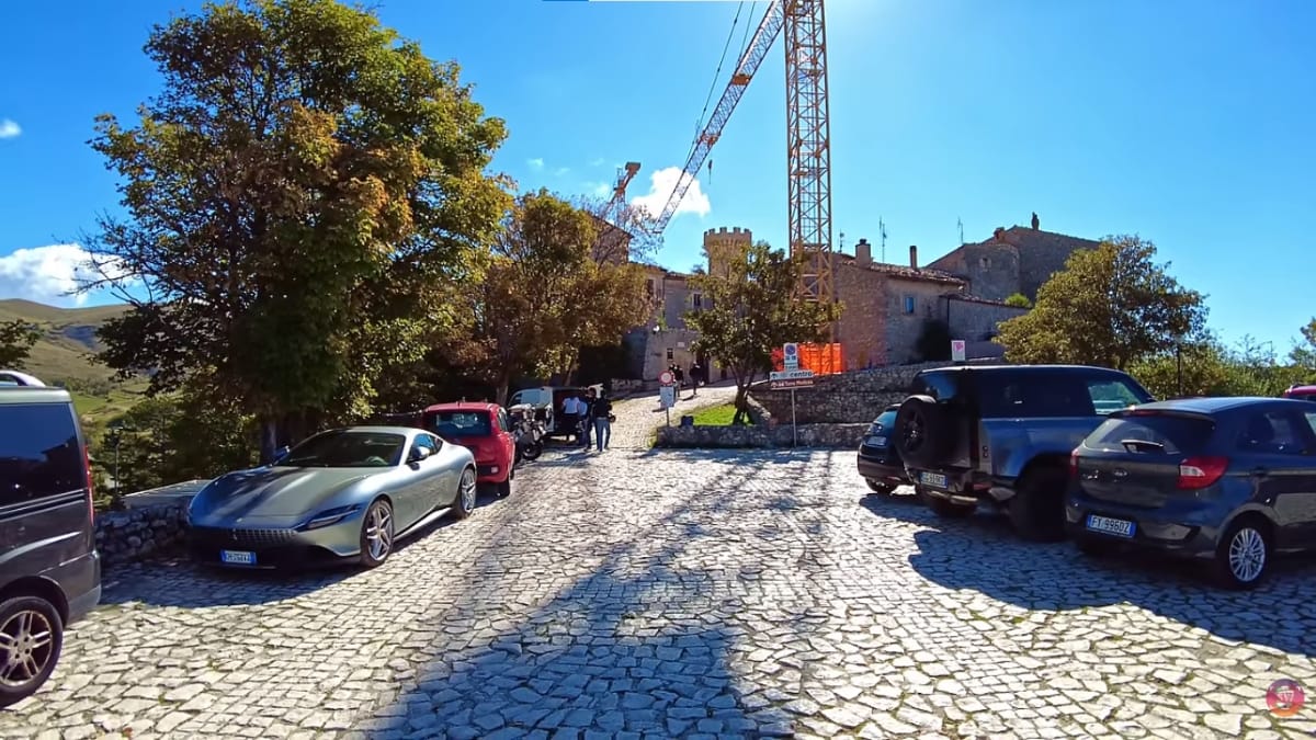 Pátio pavimentado de pedra com vários carros modernos estacionados, árvores e edifícios de pedra ao fundo, sob um céu ensolarado.