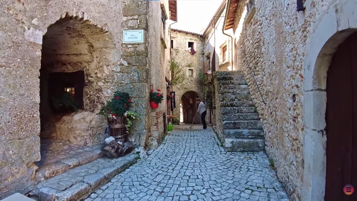 Ruela estreita de pedras cercada por paredes de edifícios antigos de pedra, com vasos de flores vermelhas pendurados e uma pessoa limpando o chão ao fundo.