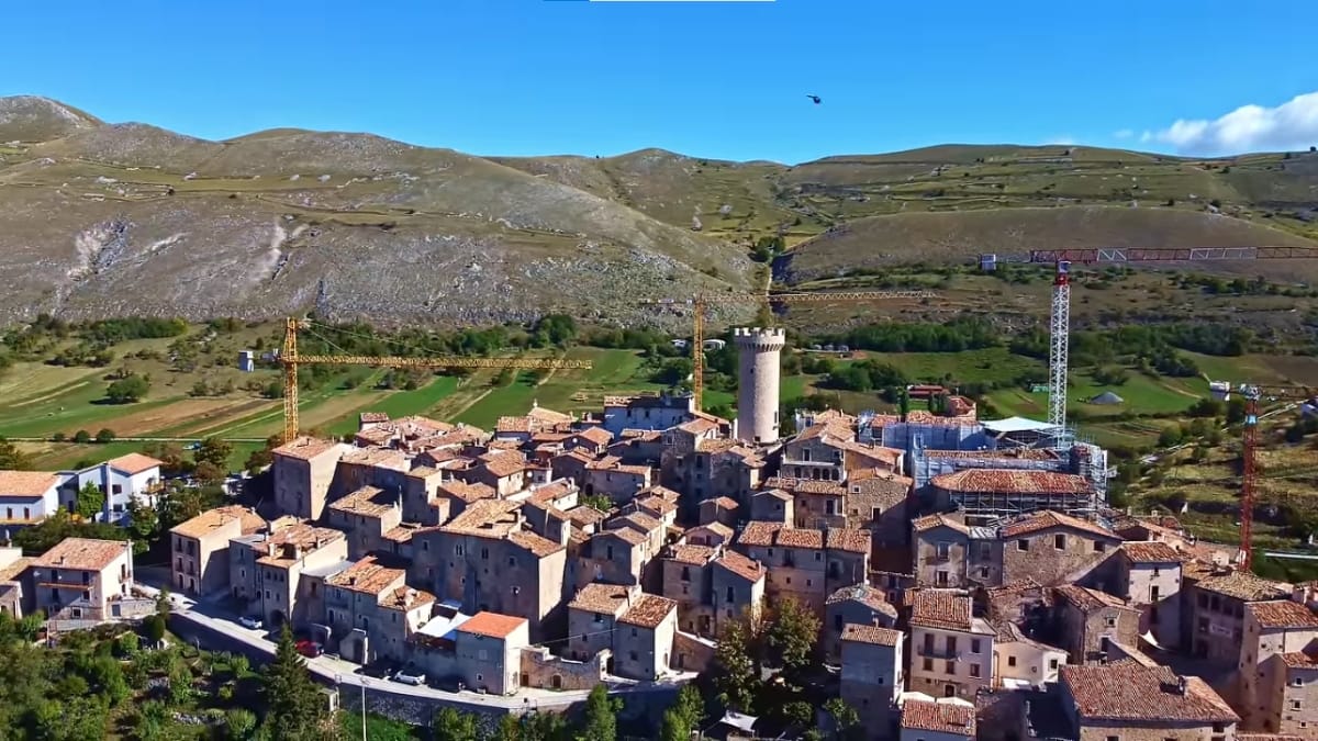 Vista aérea de uma antiga vila medieval de pedra no topo de uma colina, com montanhas verdes e áridas ao fundo sob um céu azul.