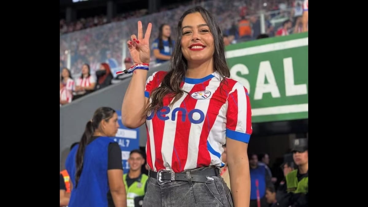Larissa Riquelme em um estádio de futebol lotado, vestindo uma camisa moderna da seleção paraguaia e calça jeans com cinto largo. Ela sorri e faz o sinal de "V" de vitória com os dedos da mão direita elevada. Ao fundo, placas do estádio e torcedores.