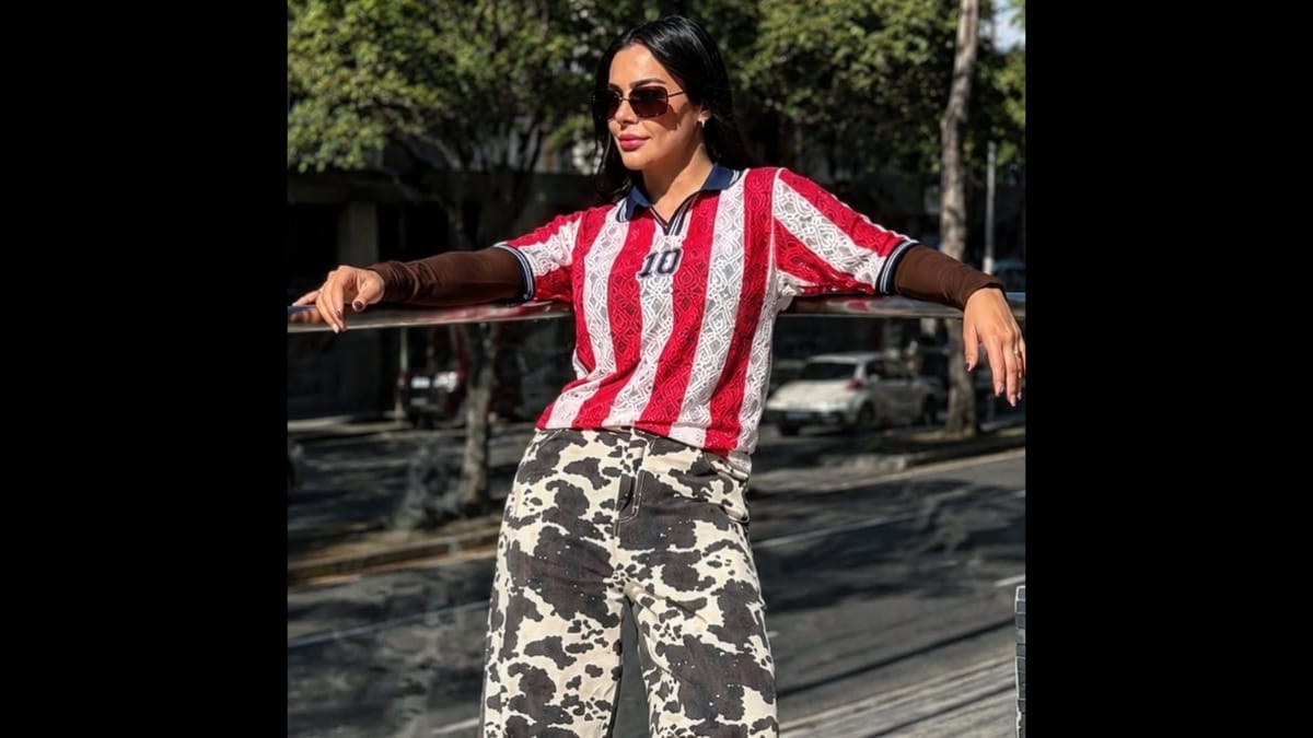 Larissa Riquelme posa ao ar livre encostada em um corrimão de metal na rua. Ela usa óculos escuros, uma camisa estilizada de futebol do Paraguai com o número 10 e uma calça comprida com estampa de vaquinha (malhada em preto e branco).