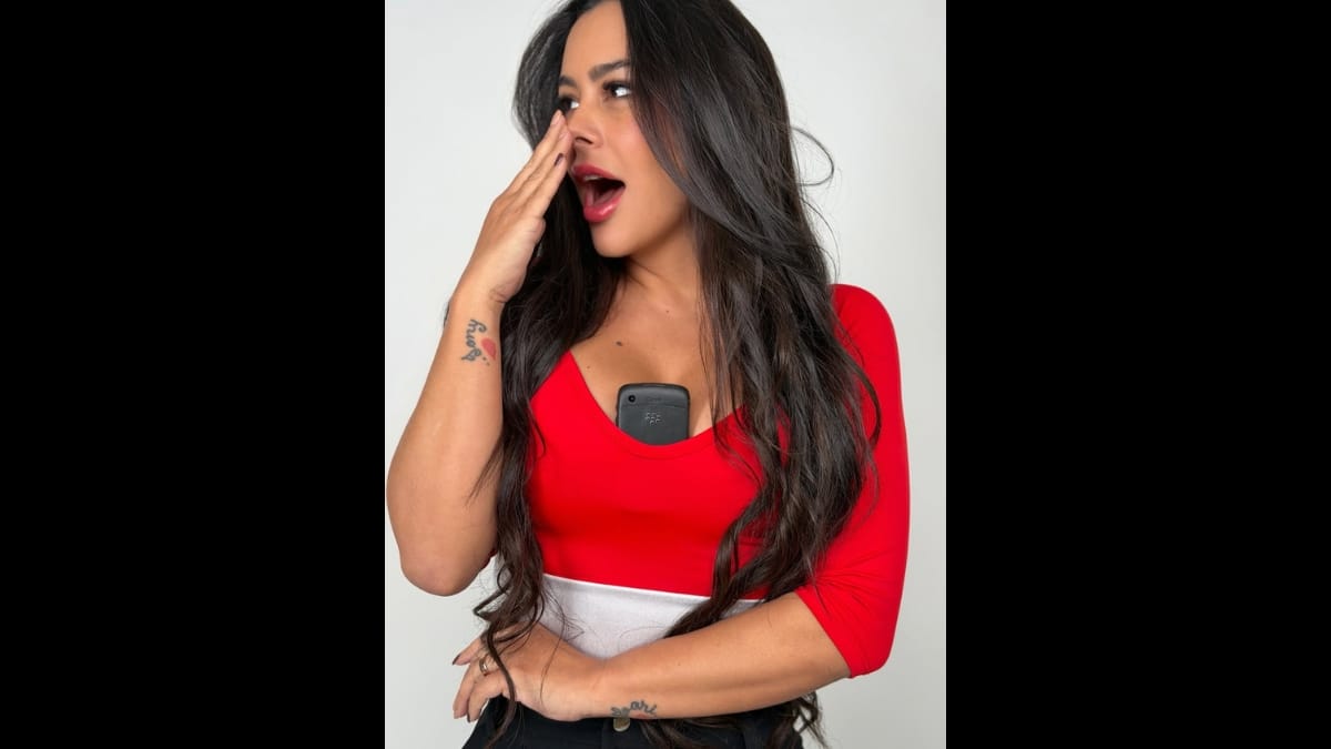 Larissa Riquelme em pose expressiva de estúdio, olhando para o lado com a boca ligeiramente aberta em sinal de surpresa, com uma das mãos perto do rosto. Ela veste a blusa temática do Paraguai com o celular no decote.