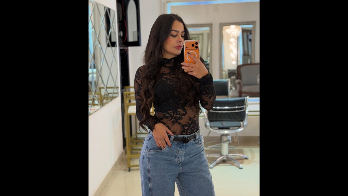 Selfie no espelho tirada por Larissa Riquelme em um salão de beleza ou camarim. Ela veste uma blusa preta de renda transparente de gola alta e calça jeans azul, segurando um smartphone moderno com capinha laranja.