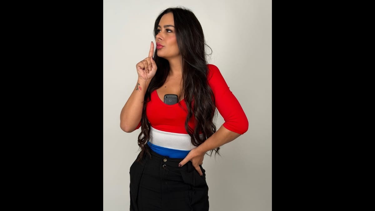 Larissa Riquelme em perfil parcial, vestindo uma blusa justa com as cores da bandeira do Paraguai (vermelho, branco e azul) e calça preta. Ela tem um celular antigo preto alojado no decote e faz um gesto de silêncio com o dedo indicador sobre os lábios.