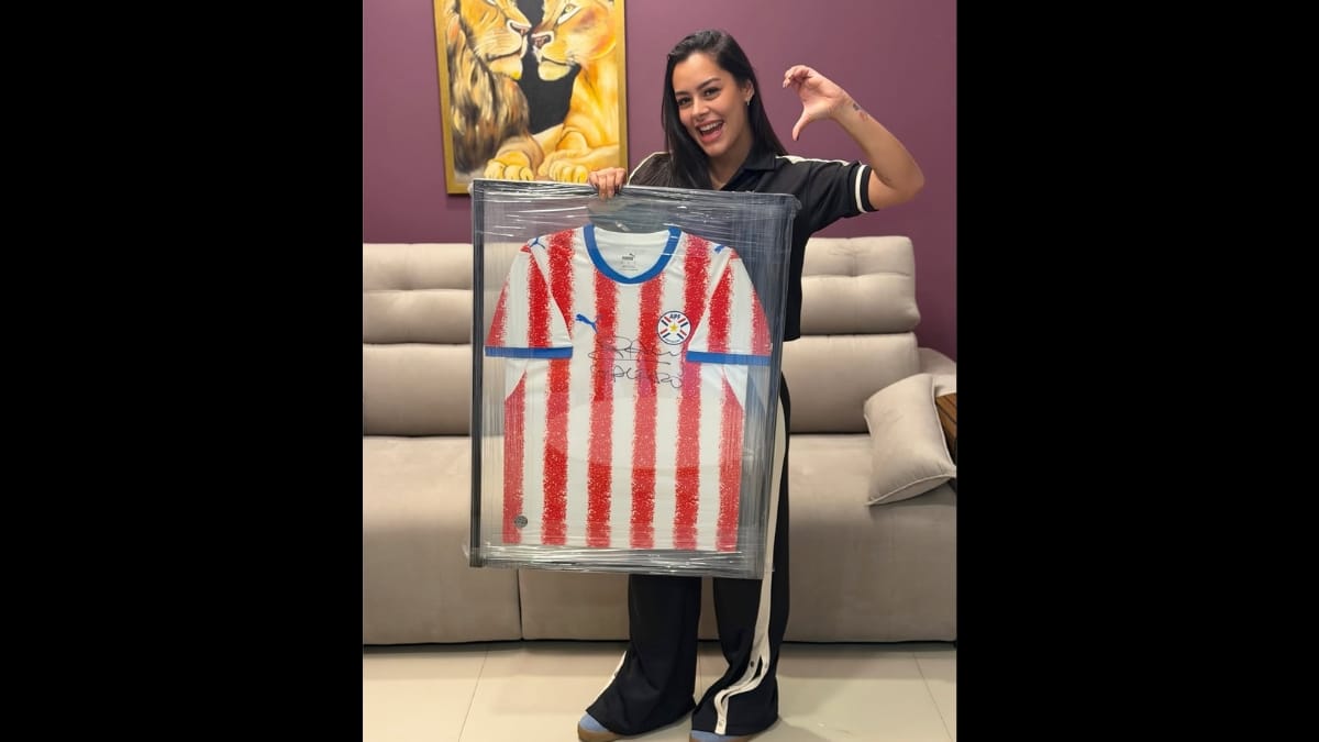 Larissa Riquelme posa sorridente segurando o mesmo quadro com a camisa do Paraguai, mas desta vez faz metade de um coração com a mão esquerda elevada na altura da cabeça.