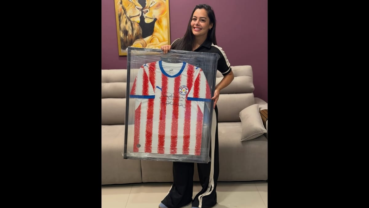 Mulher sorridente de cabelos longos e escuros, vestindo conjunto esportivo preto com listras brancas, segura orgulhosamente à sua frente uma camisa oficial da seleção do Paraguai (listrada em vermelho e branco) emoldurada e protegida por plástico. Ao fundo, uma parede roxa e um quadro com leões.