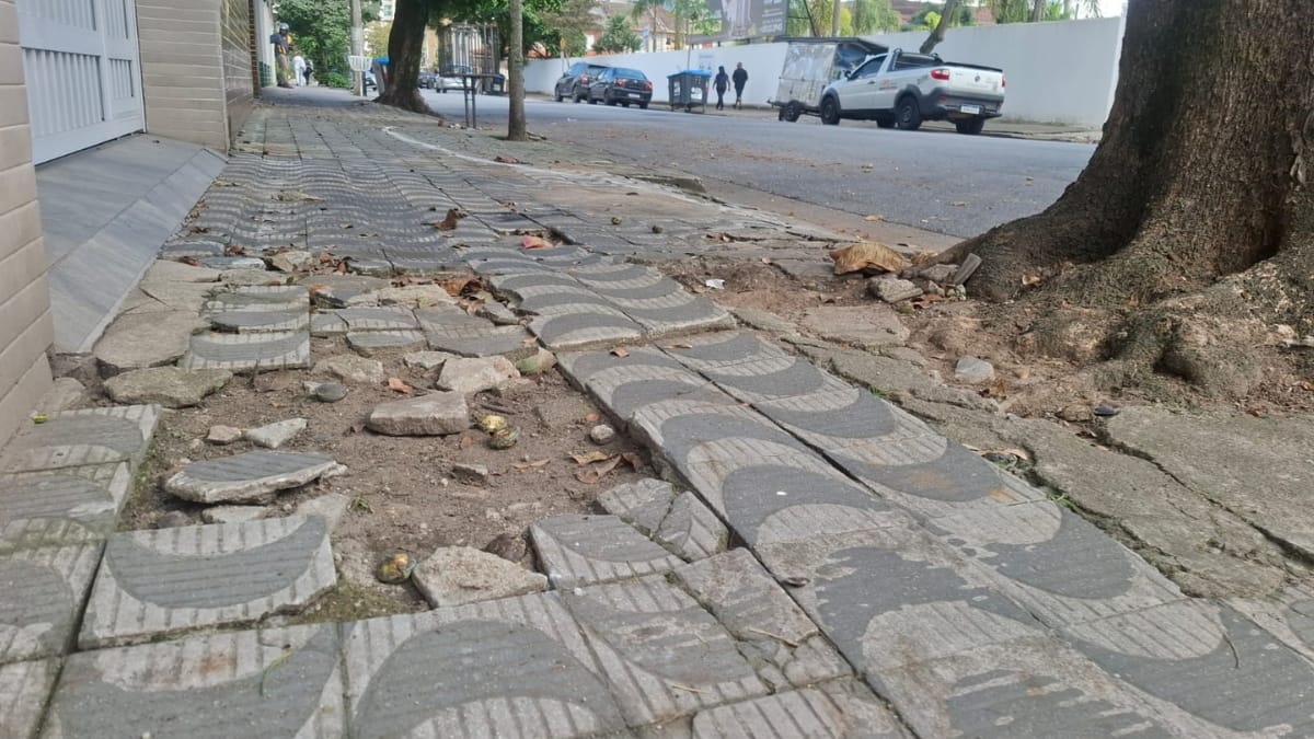 Uma foto em ângulo baixo e perspectiva profunda de uma calçada de Santos com padrão de ondas. Raízes grossas de uma árvore à direita levantaram e quebraram a calçada de azulejos, espalhando detritos. O resto da calçada é irregular e perigoso. Carros estacionados estão à direita.